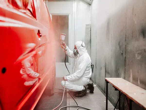 Ein Lackierer trägt in einer Werkstatt Schutzkleidung und lackiert ein rotes Auto professionell.